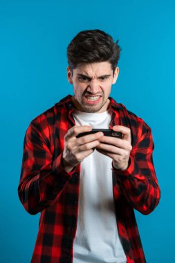 Mavi stüdyo duvarında akıllı telefondan oyun oynayan yakışıklı bir adam. Modern teknolojiyi kullanarak - uygulamalar, sosyal ağlar.