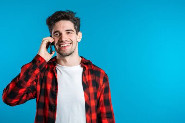 Parlak renkli giyinmiş genç adam mavi arka planda cep telefonuyla konuşuyor. Modayı takip eden adam konuşuyor. Akıllı telefon, teknoloji konsepti.