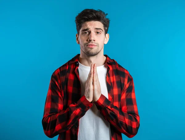 Bonito homem de camisa vermelha rezando sobre fundo azul. Europeu raça cara implorando alguém . — Fotografia de Stock