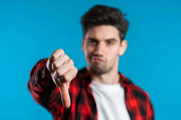 Homem bonito jovem em pé no fundo do estúdio azul expressando descontentamento e mostrando o polegar para baixo gesto na câmera. Retrato de cara com sinal de antipatia — Fotografia de Stock