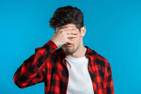 Young guy covers his face with hands from fatal disappointment, failure. Depressed lost man in red plaid shirt. Drama, emotions concept.