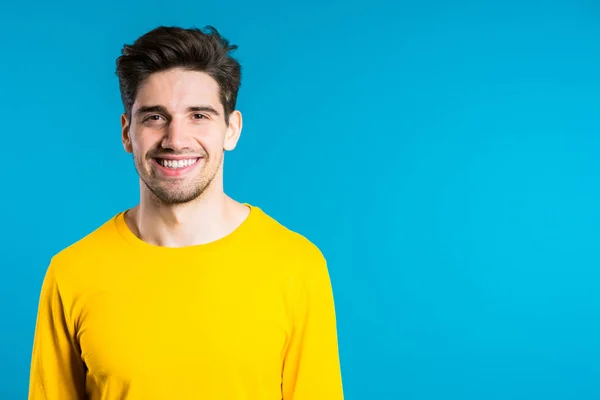 Homem europeu bonito com penteado na moda em amarelo desgaste no fundo do estúdio azul. Espaço para cópia. cara alegre sorrindo e olhando para a câmera . — Fotografia de Stock