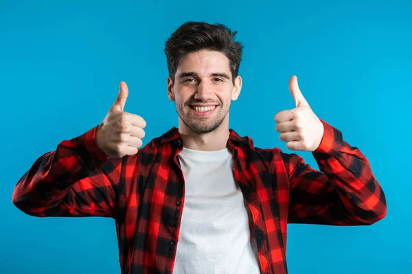 Positive young man smiles to camera. Hipster guy showing thumb up sign over blue background. Winner. Success. Body language. — Stock Photo, Image