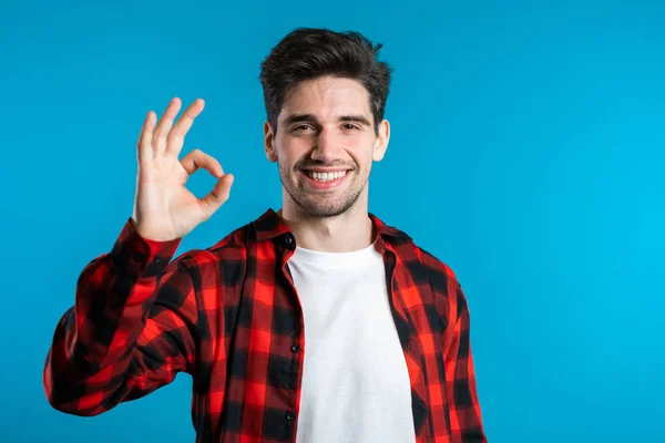 Handsome young man in red plaid shirt smiles to camera. Hipster guy showing OK sign over blue background. Winner. Success. Body language. — Stock Photo, Image