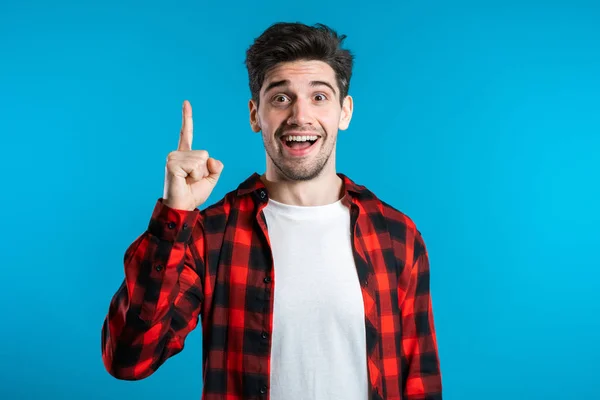 Retrato del joven hombre pensante en rojo teniendo idea momento señalando el dedo hacia arriba en el fondo del estudio azul. Sonriendo feliz estudiante chico mostrando eureka gesto . — Foto de Stock