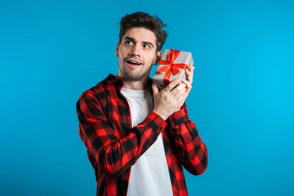 Handsome man holding gift box on blue studio background smiles to camera. Happy european guy received present and interested in whats inside. — Stock Photo, Image