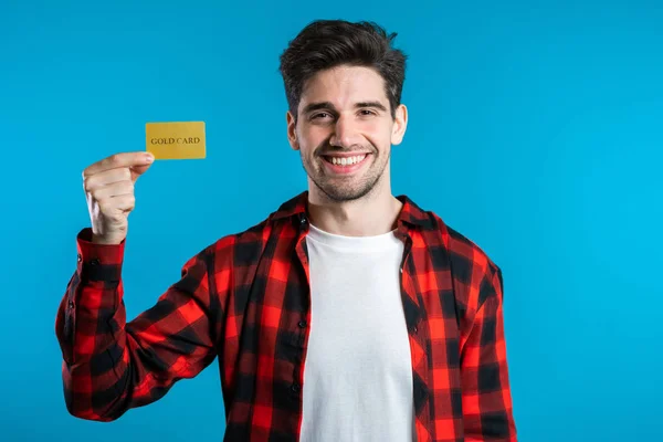 Successful man in plaid shirt showing unlimited gold credit card on blue studio background. Student, money concept — Stock Photo, Image