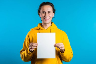 Beyaz A4 kağıt poster tutan yakışıklı bir adam. Uzayı kopyala Mavi arka planda sarı kapüşonlu gülümseyen hippi..