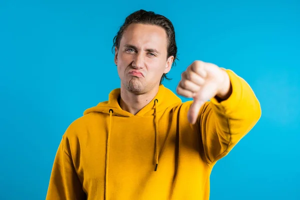 Young handsome man standing on blue studio background expressing discontent and showing thumb down gesture at camera. Portrait of guy with sign of dislike — Stock Photo, Image