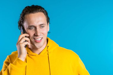 Parlak sarı elbiseli genç adam mavi arka planda cep telefonuyla konuşuyor. Modayı takip eden adam konuşuyor. Akıllı telefon, teknoloji konsepti.