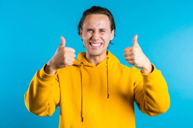 Olumlu genç adam kameraya gülümsüyor. Hippi adam mavi arka planda baş parmak işareti gösteriyor. Kazandım. Başarı. Vücut dili.