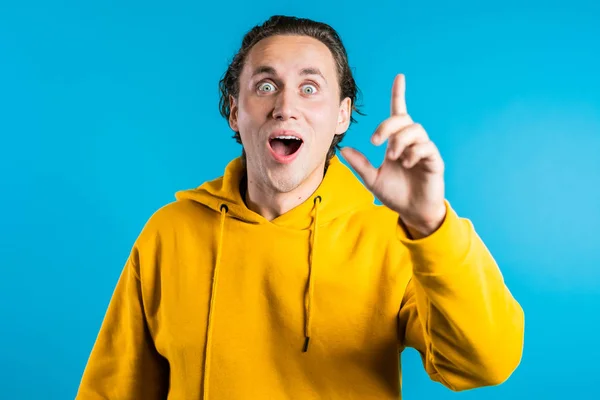 Portrait of young thinking pondering man in yellow having idea moment pointing finger up on blue studio background. Smiling happy student guy showing eureka gesture. — Stock Photo, Image