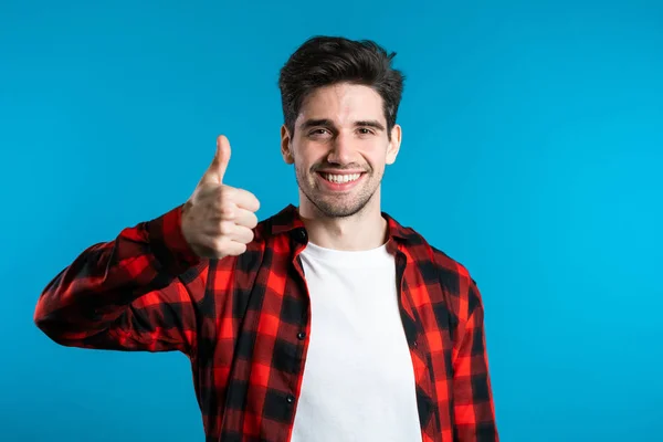 一个积极向上的年轻人对着摄像机笑. 希斯特的家伙用大拇指在蓝色背景上做记号. 赢了 成功。 身体语言. — 图库照片