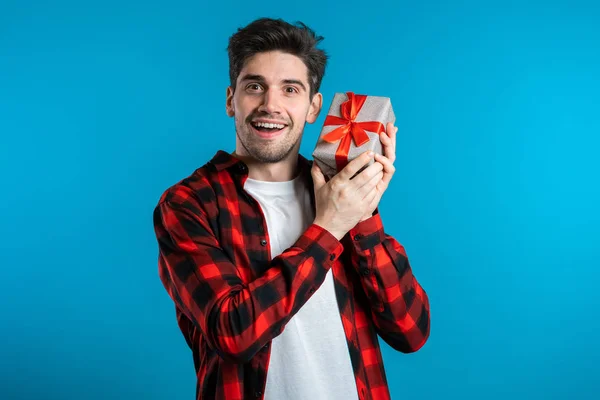 Bonito homem segurando caixa de presente no fundo do estúdio azul sorri para a câmera. Cara europeia feliz recebeu presente e interessado no que está dentro . — Fotografia de Stock