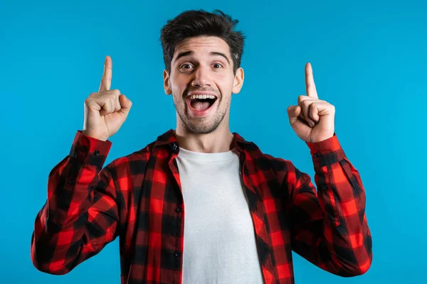 Porträt eines nachdenklichen jungen Mannes in Rot, der im Ideenmoment mit dem Finger auf blauem Studiohintergrund zeigt. Glücklich lächelnder Student zeigt Eureka-Geste. — Stockfoto