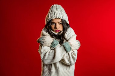 Warmly dressed woman shaking from low temperature in knitted hat, sweater and mittens. She freezing and shows brrr how cold it is in winter. Studio red background. clipart