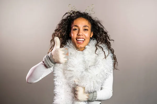 Mujer afroamericana joven en manoplas de punto suéter cálido y cuernos de ciervo sonriendo y mostrando pulgares hacia arriba signo sobre fondo gris estudio. Chica en buen humor de Navidad de invierno . — Foto de Stock