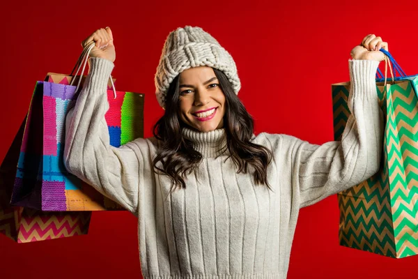 Glückliche junge Frau mit bunten Papiertüten nach dem Einkauf isoliert auf rotem Studiohintergrund. Winterschlussverkauf, Einkäufe, Geld für Geschenkkonzept ausgeben. — Stockfoto