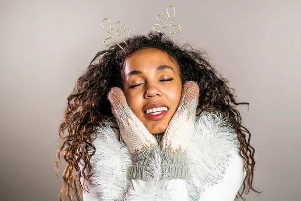 Retrato de mujer afroamericana en mitones de punto, cuernos de ciervo y suéter cálido sonriendo sobre fondo gris del estudio. Chica en buen humor de Navidad de invierno . — Foto de Stock