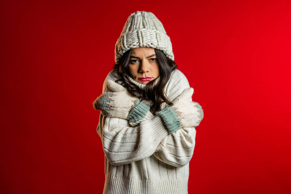 Warmly dressed woman shaking from low temperature in knitted hat, sweater and mittens. She freezing and shows brrr how cold it is in winter. Studio red background.