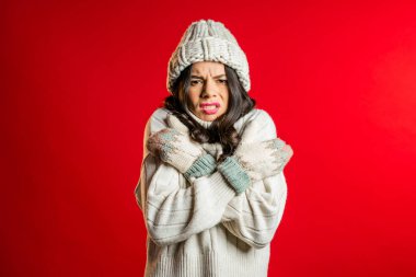 Warmly dressed woman shaking from low temperature in knitted hat, sweater and mittens. She freezing and shows brrr how cold it is in winter. Studio red background. clipart