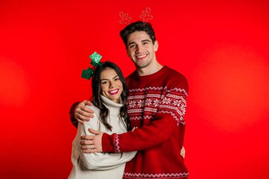 Kırmızı stüdyo arka planında Noel süsleri takmış genç bir çiftin portresi. Aşk, tatiller, mutluluk konsepti.