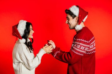 Noel Baba şapkalı genç çift, kırmızı stüdyo geçmişinde yüzüğü olan sevgilisine evlenme teklif ediyor. Aşk, tatiller, mutluluk konsepti.