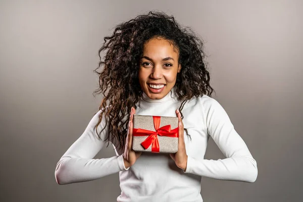 Giovane ragazza razza mista sorridente e in possesso di scatola regalo su sfondo grigio studio. Carino il ritratto di una donna afroamericana. L'umore di Natale — Foto Stock