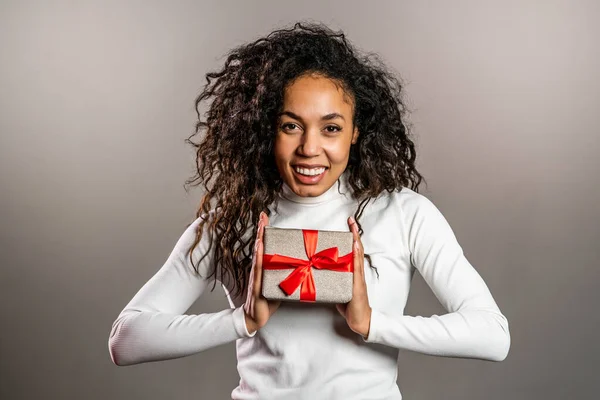 Ung blandad ras flicka ler och håller presentförpackning på grå studio bakgrund. Gulligt porträtt av en afrikansk amerikansk kvinna. Julstämning — Stockfoto