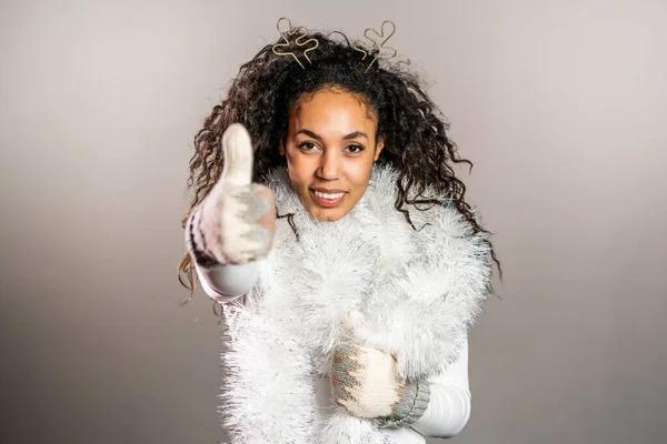 Молода афроамериканська жінка в в'язаній рукавичці теплого светра і роги оленів посміхаються і показують пальці вгору на сірому студійному фоні. Дівчинка в хорошому зимовому настрої. — стокове фото