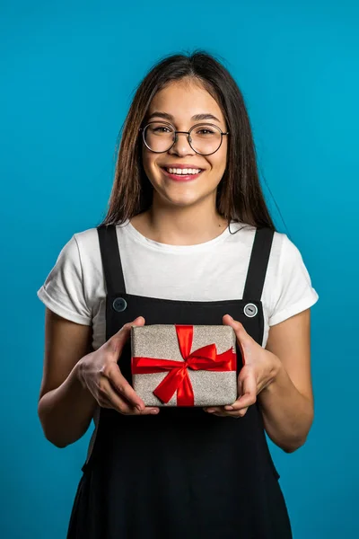 Νεαρό χαριτωμένο κορίτσι χαμογελά και κρατώντας κουτί δώρου σε μπλε φόντο στούντιο. Χαριτωμένο πορτρέτο φοιτήτριας. Χριστουγεννιάτικη διάθεση. — Φωτογραφία Αρχείου
