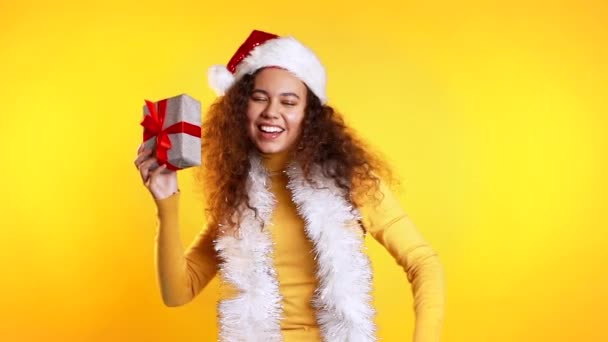 年轻漂亮的非洲女人带着礼品盒在黄色工作室的背景下微笑和跳舞。 穿着毛衣的卷发女孩。 圣诞气氛. — 图库视频影像