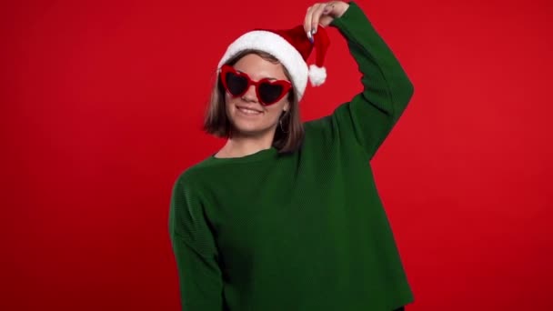 Junge hübsche Frau lächelt und tanzt auf rotem Studiohintergrund. Mädchen mit Weihnachtsmütze. Neujahrsstimmung. Zeitlupe. — Stockvideo