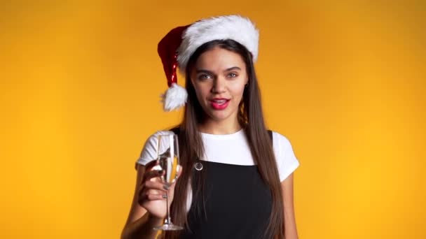 Ung söt flicka ler och håller glas champagne eller vin på gul studio bakgrund. Kvinnan i tomteluva. Nytt år humör — Stockvideo