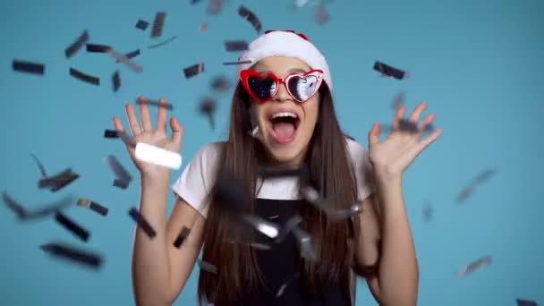 Chica emocionada con Santa sombrero saltando, aplaudiendo, divirtiéndose, se regocija por la lluvia de confeti en el estudio azul. Concepto de Navidad, Año Nuevo, felicidad, fiesta, ganar . — Vídeo de stock