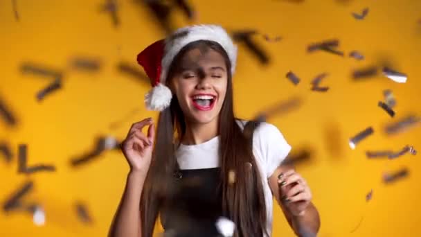 Захоплена дівчиною з Санта-Катом, що стрибає, аплодує, розважається через дощ конфетті в жовтій студії. Концепція Різдва, Нового року, щастя, вечірки, перемоги. — стокове відео