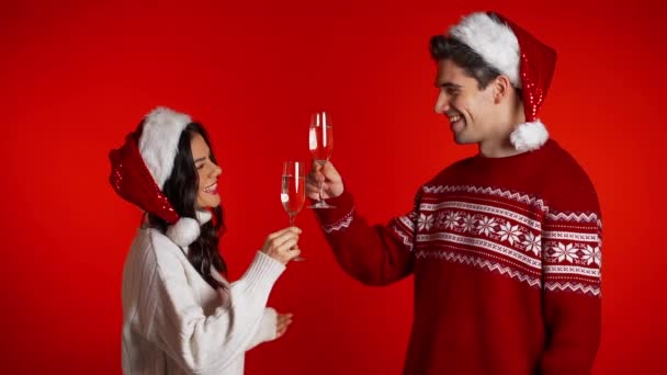 Portré fiatal pár karácsonyi Mikulás kalap iszik pezsgőt piros stúdió háttér. Szerelem, ünnepek, boldogság koncepció — Stock videók