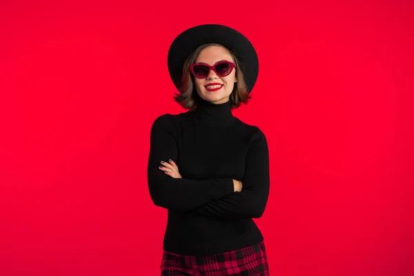 Ritratto di giovane bella donna su sfondo rosso in studio. Ragazza alla moda hipster in cappello nero, occhiali da sole e mini gonna a quadri. Signora sorridente . — Foto Stock