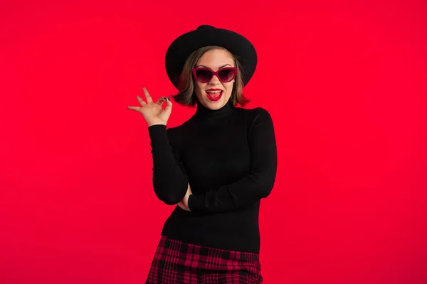 Porträt einer jungen hübschen Frau mit Brille, die im Atelier ein Ok-Zeichen auf rotem Hintergrund zeigt. trendiges Hipster-Mädchen mit schwarzem Hut und kariertem Minirock. Lächelnde Dame. — Stockfoto