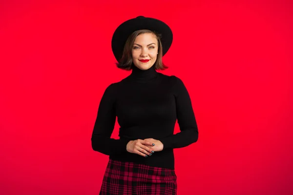 Portrait of young pretty woman on red background in studio. Trendy hipster girl in black hat and plaid mini skirt. Smiling lady. — Stock Photo, Image