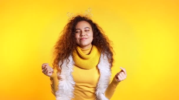 Mujer joven bastante mestiza sonriendo y bailando sobre fondo amarillo estudio. Chica con peinado rizado y con corona de Navidad. El humor de Año Nuevo . — Vídeos de Stock