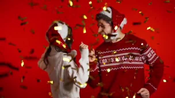 Pareja excitada Santa Claus sombreros bailando, aplaudiendo, divirtiéndose, se regocija por la lluvia de confeti en el estudio rojo. Concepto de Navidad, Año Nuevo, felicidad, fiesta, ganar . — Vídeos de Stock