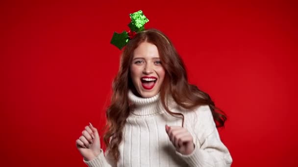 Fiatal csinos nő mosolyog és táncol a vörös stúdió hátterében. Karácsonyi koszorús lány. Újévi hangulat. Lassú mozgás.. — Stock videók
