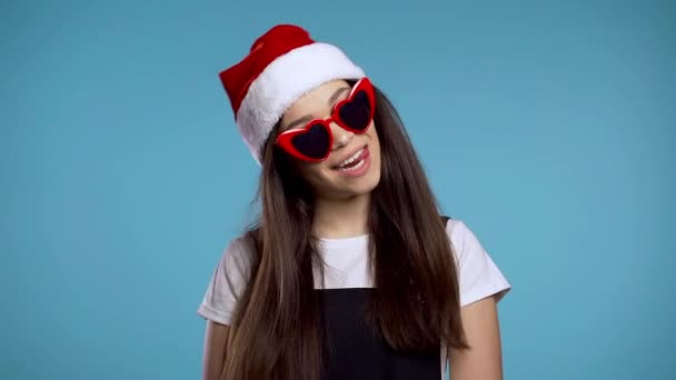 Attraktives Mädchen mit Weihnachtsmütze und herzförmiger Sonnenbrille, das Weihnachten oder Neujahr auf blauem Hintergrund feiert. glückliche hübsche Frau, die lächelt und Spaß hat. — Stockvideo