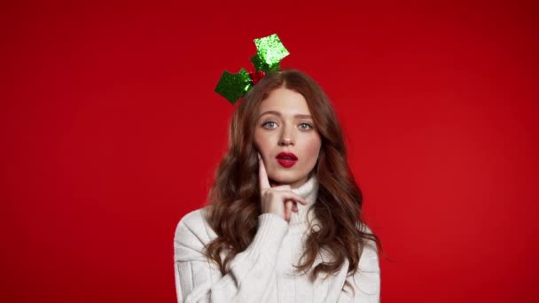 Tänkande kvinna i julkrans titta upp och runt på röd bakgrund. Oroade kontemplativa ansiktsuttryck. Vacker ung modell med vacker frisyr. — Stockvideo