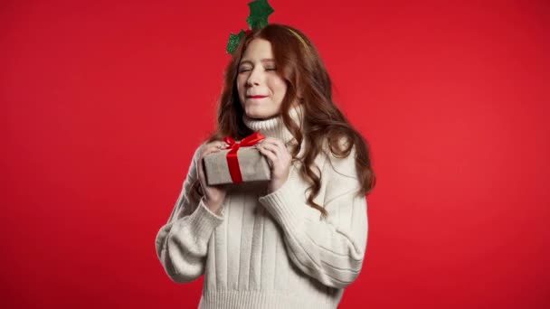 若いかわいい女の子は笑顔と赤いスタジオの背景にプレゼントボックスを保持します。クリスマスの頭の花輪の女性。新年の気分. — ストック動画