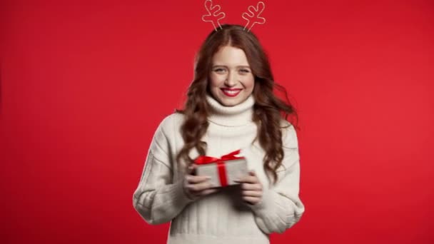 Menina bonito jovem sorrindo e segurando caixa de presente no fundo do estúdio vermelho. Mulher na grinalda de cabeça de Natal - chifres de veado. Modo de ano novo . — Vídeo de Stock