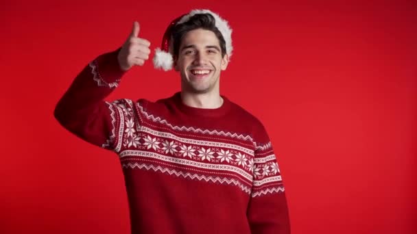 Joyeux jeune homme au chapeau de Père Noël montrant pouces vers le haut geste isolé sur fond de studio rouge. Concept vacances d'hiver . — Video