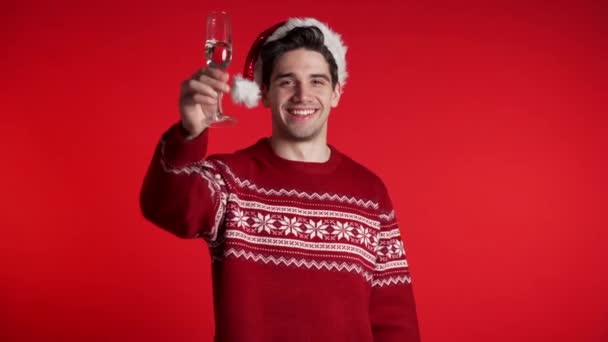 Bonito homem sorrindo e segurando copo de champanhe ou vinho no fundo do estúdio vermelho. Um tipo de chapéu de Pai Natal. Ano Novo festa 2020 humor — Vídeo de Stock