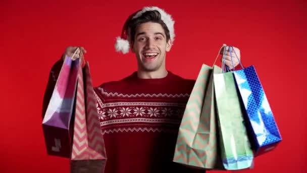 Hombre joven guapo en jersey de Navidad y sombrero de Santa sostiene bolsas de papel de compras en el fondo del estudio rojo. Chico compró regalos en ventas con descuentos . — Vídeo de stock